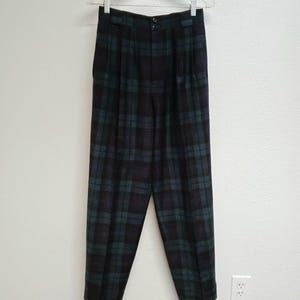 May include: Dark green and black plaid trousers hanging on a clear hanger. The pants have a high waist, pleats, and a button closure. The plaid pattern is a classic design.