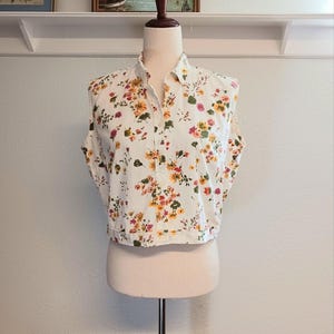 May include: A sleeveless, button-down shirt on a mannequin. The shirt is white with a colorful floral pattern featuring yellow, orange, pink, and green flowers. The shirt has a collared neckline and a cropped hemline.