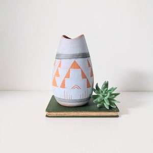 May include: A light blue vase with orange geometric patterns and gray accents. The vase is displayed on a green book with a small succulent plant next to it. The vase has a unique shape and design.
