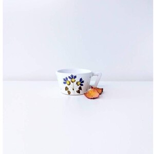 May include: White ceramic teacup with a floral design featuring blue and yellow flowers and brown leaves. The teacup has a handle and is set on a white surface. Dried orange petals are next to the cup.