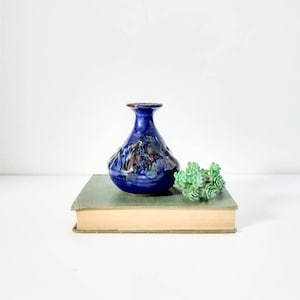 May include: A small, blue ceramic vase with a textured surface and a brown rim. The vase sits on a vintage book with a green cover, next to a small, green succulent plant. The vase has a unique shape.