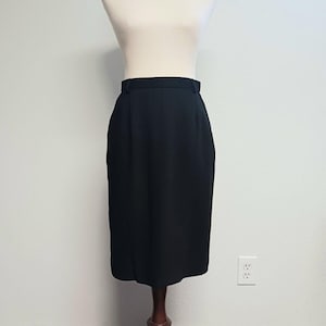 May include: A black pencil skirt displayed on a mannequin. The skirt is knee-length with a straight silhouette and a waistband. The fabric appears to be a smooth, solid color. The skirt is displayed on a wooden stand against a white wall.