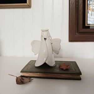 May include: A white ceramic angel figurine with outstretched wings, standing on a vintage green book. A dried leaf rests on the book, and another is nearby. The angel has a halo and flowing robes.