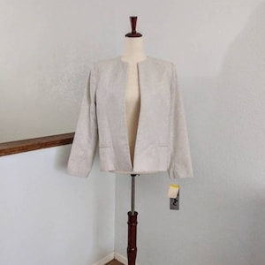 May include: A light beige blazer with thin vertical stripes, displayed on a wooden dress form. The jacket has long sleeves, a collarless design, and two front pockets. A tag hangs from the jacket.