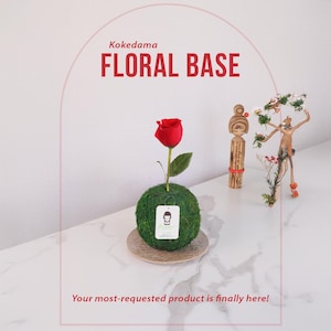 May include: A single red rose with green leaves is displayed in a kokedama floral base. The base is a sphere of moss with a small brown plate underneath. The text "Kokedama FLORAL BASE" is displayed above the image.