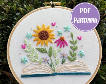 May include: Embroidered hoop art featuring a book with a sunflower, pink and purple flowers, and green leaves. The book is open and the flowers are growing out of it. The background is white and the hoop is made of wood.