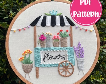 Peut inclure: Un cerceau de broderie blanc avec un motif de charrette à fleurs colorée. La charrette a une auvent rayé noir et blanc et dit "flowers" sur le devant. Le motif comprend une variété de fleurs, de pots et une roue.
