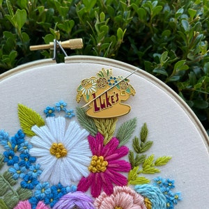 May include: A gold-coloured enamel pin featuring a coffee cup with the text "Luke's" and a floral design. The pin is attached to a white fabric with embroidered flowers in shades of pink, purple, blue, and green.