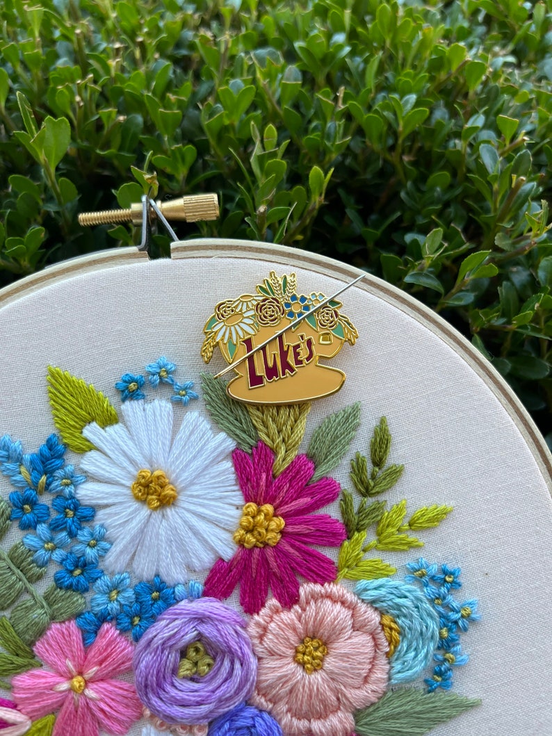 May include: A gold-coloured enamel pin featuring a coffee cup with the text "Luke's" and a floral design. The pin is attached to a white fabric with embroidered flowers in shades of pink, purple, blue, and green.