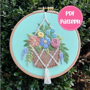 Puede incluir: Un aro de bordado cosido a mano con una maceta colgante de macramé llena de flores coloridas. El aro es de madera y tiene un fondo de tela verde claro. La maceta es de fieltro marrón y tiene una borla blanca.