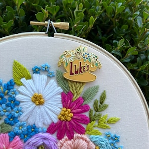 May include: A white embroidery hoop featuring a colourful floral design. A gold enamel pin with the text "Luke's" is attached to the hoop.