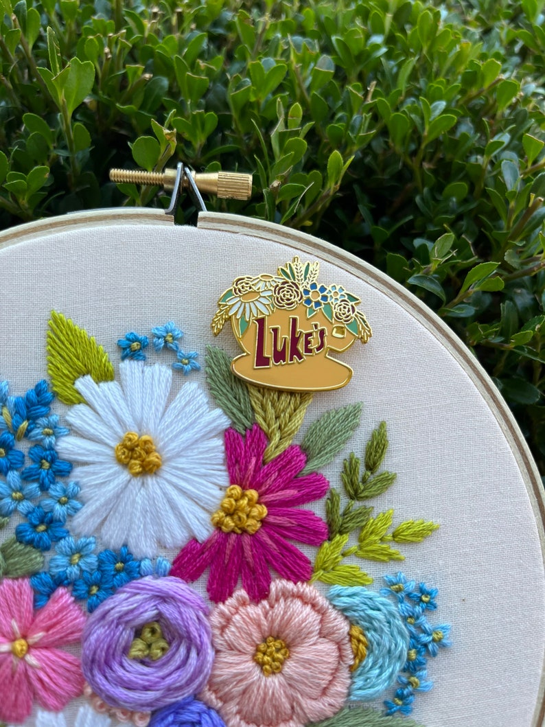May include: A white embroidery hoop featuring a colourful floral design. A gold enamel pin with the text "Luke's" is attached to the hoop.