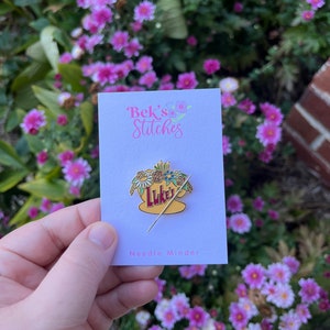 May include: A gold needle minder featuring a yellow mug with the text "Luke's" and a floral arrangement. The needle minder is on a white card with the text "Bek's Stitches" and "Needle Minder".