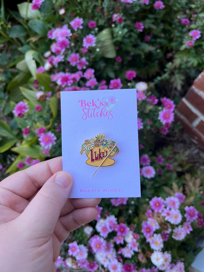 May include: A gold needle minder featuring a yellow mug with the text "Luke's" and a floral arrangement. The needle minder is on a white card with the text "Bek's Stitches" and "Needle Minder".
