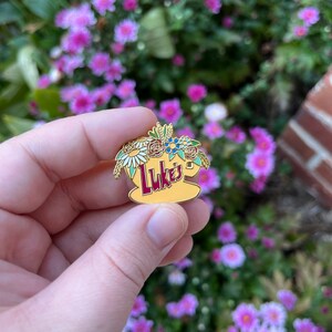 May include: A gold-coloured enamel pin featuring a yellow coffee cup with the word "Luke's" in red lettering. The cup is surrounded by a floral arrangement of white, red, blue, and green flowers.