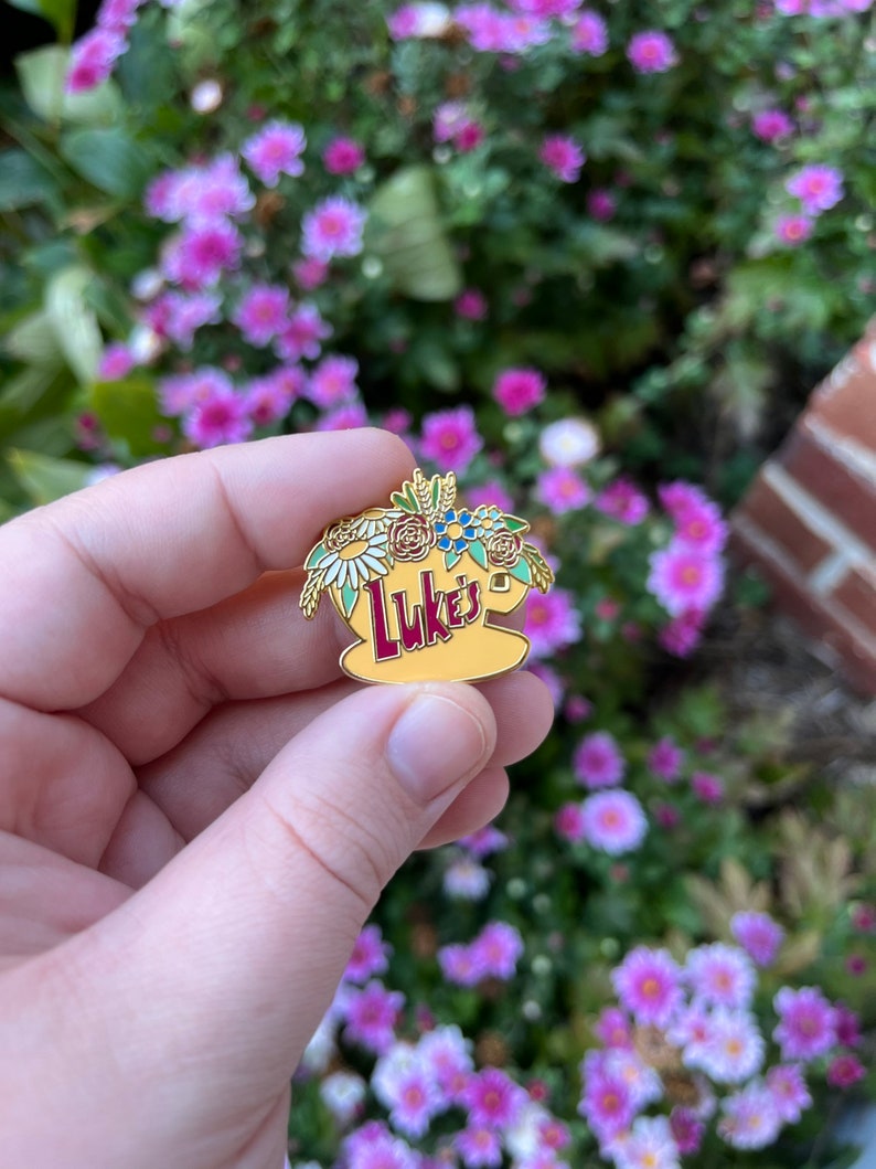 May include: A gold-coloured enamel pin featuring a yellow coffee cup with the word "Luke's" in red lettering. The cup is surrounded by a floral arrangement of white, red, blue, and green flowers.