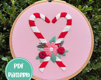 Peut inclure: Art de broderie en forme de cercle représentant deux cannes en sucre formant un cœur avec un arrangement floral au centre. Le cercle est rose avec un cadre en bois. Les cannes en sucre sont rouges et blanches, et les fleurs sont roses, vertes et rouges.