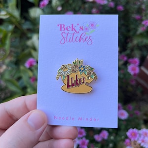 May include: A gold-coloured needle minder with a floral design and the text "Luke's" in red. The needle minder is on a white card with the text "Bek's Stitches" and "Needle Minder".