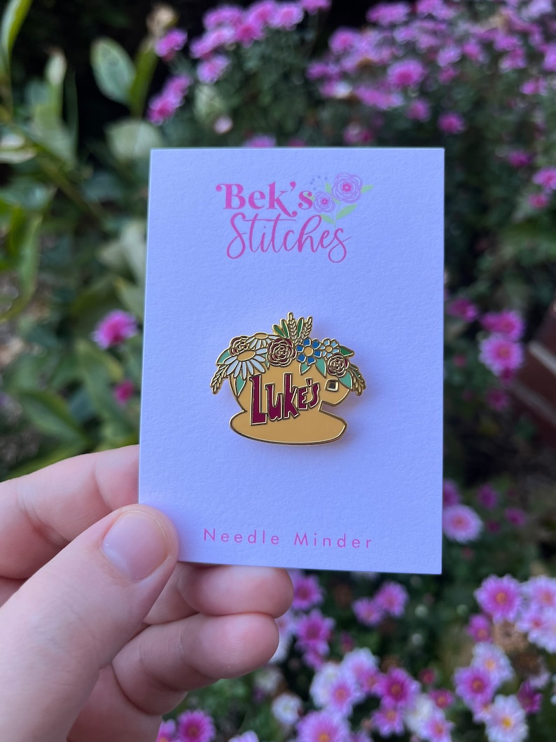 May include: A gold-coloured needle minder with a floral design and the text "Luke's" in red. The needle minder is on a white card with the text "Bek's Stitches" and "Needle Minder".