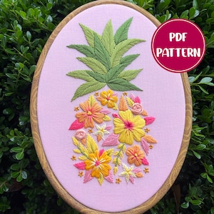 Puede incluir: Diseño de piña bordado con flores rosas, amarillas y verdes sobre un fondo de tela rosa en un aro de bordado de madera. PATRÓN PDF.