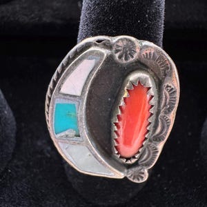 May include: Silver ring with a central red coral stone, surrounded by a crescent of white and turquoise stones. The ring has an ornate, engraved border and a dark background.
