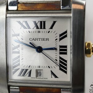 May include: Close-up of a Cartier Tank watch. The watch has a rectangular silver dial with Roman numeral hour markers, a date window at the 3 o'clock position, and a blue steel hands. The watch case is made of stainless steel with a brown leather strap. The watch face features a guilloché pattern and the Cartier logo is visible at the center. The text 'SWISS MADE' is printed at the bottom of the dial.