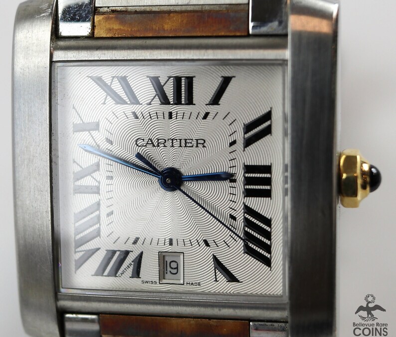May include: Close-up of a Cartier Tank watch. The watch has a rectangular silver dial with Roman numeral hour markers, a date window at the 3 o'clock position, and a blue steel hands. The watch case is made of stainless steel with a brown leather strap. The watch face features a guilloché pattern and the Cartier logo is visible at the center. The text 'SWISS MADE' is printed at the bottom of the dial.