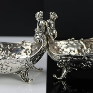 Puede incluir: Un par de platos de servir de plata con detalles ornamentados y un diseño de querubín en las asas. Los platos están sobre un fondo blanco y negro.
