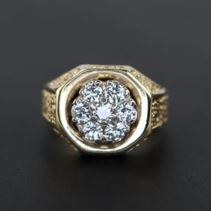 Può includere: Un anello d'oro con un grande diamante centrale rotondo e chiaro circondato da diamanti più piccoli. L'anello ha una fascia testurizzata.