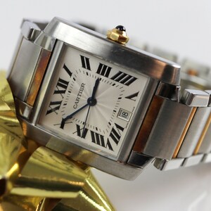 May include: Close-up of a Cartier Tank watch. The watch has a square, silver face with Roman numerals and a date window. The case is stainless steel and two-toned with gold accents. The bracelet is a multi-link design with alternating steel and gold links. The watch is displayed on a gold ribbon.