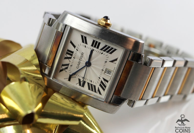 May include: Close-up of a Cartier Tank watch. The watch has a square, silver face with Roman numerals and a date window. The case is stainless steel and two-toned with gold accents. The bracelet is a multi-link design with alternating steel and gold links. The watch is displayed on a gold ribbon.