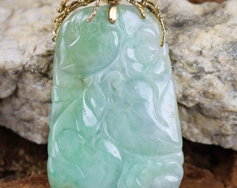 May include: A green jade pendant with intricate floral carvings, set in a gold frame with a delicate design. The pendant is a rectangular shape with rounded corners and is approximately 2 inches in length.