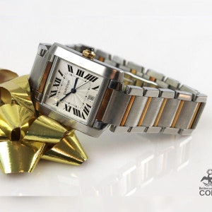 May include: A Cartier Tank watch with a stainless steel and gold-tone bracelet. The watch has a rectangular white dial with Roman numeral hour markers and a date window. The watch is displayed on a gold ribbon.  The brand name 'CARTIER' is visible on the dial.