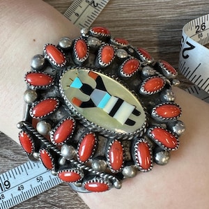 Puede incluir: Una pulsera de plata con una incrustación ovalada central que presenta una figura geométrica en negro, blanco, turquesa y gris. La pulsera está adornada con coral rojo y cuentas de plata, mostrando un diseño del suroeste.