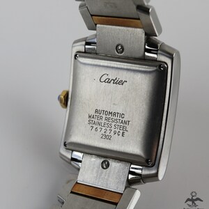 May include: Close-up of the back of a Cartier watch. The stainless steel case back is engraved with 'Cartier', 'Automatic', 'Water Resistant', 'Stainless Steel', '767279CE', and '2302'. The watch features a two-tone band with stainless steel and gold accents. The watch is a classic design with a square face and a polished finish.
