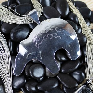 May include: A polished silver pendant in the shape of a bear, with etched details on its back. The pendant hangs from a silver bail. The bear pendant is displayed on a bed of black stones, with strands of silver chain.