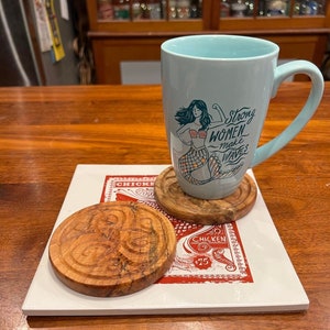May include: A light blue ceramic mug with a mermaid graphic and the text "Strong Women Make Waves". The mug is sitting on a wooden coaster with a red and white design.