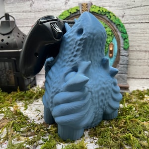 May include: A blue 3D printed dragon-like creature with a textured surface, holding a black video game controller.
