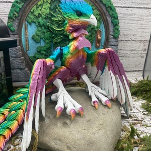 May include: A colorful fantasy bird figurine with a rainbow of colors, including blue, green, purple, and orange. The bird is perched on a gray stone. The wings are a mix of white and purple. The bird has a white beak and claws.