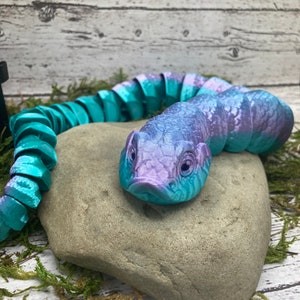 Puede incluir: Un juguete de serpiente segmentado de color verde azulado y morado con una superficie texturizada y escamosa. La serpiente está enroscada en una roca gris.