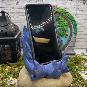 May include: A blue and gray 3D printed phone stand shaped like a dragon's head. The stand is sitting on a rock-like base.