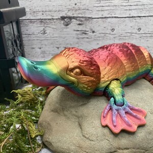 Pode incluir: Uma figura de ornitorrinco impressa em 3D com as cores do arco-íris. O brinquedo está sobre uma pedra cinzenta, com um bico detalhado, patas palmadas e um corpo texturizado. As cores variam do rosa ao amarelo e ao azul. É um artigo decorativo único.