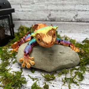 May include: A colorful, 3D printed gecko figurine with a spiky crest, perched on a gray rock. The gecko is painted in shades of orange, yellow, green, blue, and purple.