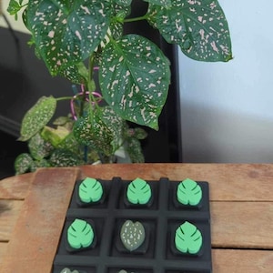 May include: A black and green tic-tac-toe game with leaf-shaped pieces. The game is on a wooden surface.