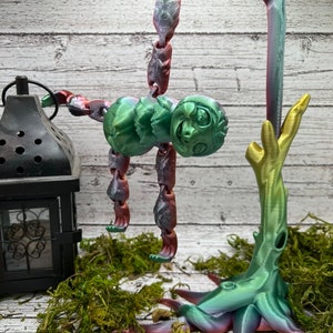 May include: A 3D printed figurine of a green and purple creature hanging from a branch. The creature has a large head with a friendly face and a long, segmented body. The branch is also 3D printed and has a gold and green iridescent finish.