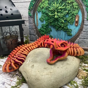 Könnte beinhalten: Eine 3D-gedruckte, bewegliche Schlange in Orange-, Rosa- und Lilatönen, die sich auf einem grauen Stein windet. Das Maul der Schlange ist geöffnet und zeigt ein rosa Inneres. Der Hintergrund umfasst eine grüne Tür mit einem Gesicht und eine schwarze Laterne.