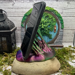 Puede incluir: Un soporte para teléfono impreso en 3D con forma de árbol con un acabado iridiscente verde, morado y rosa. El soporte está apoyado en una roca gris y un teléfono inteligente negro está apoyado contra él.