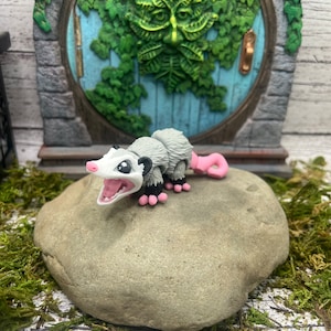 May include: A small, gray and white possum figurine with pink ears and a pink tail. The possum is standing on a gray rock with a green mossy background.