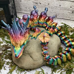 Puede incluir: Una figurita de dragón iridiscente arcoíris con cola enroscada y cabeza puntiaguda. El dragón está descansando sobre una roca gris con musgo y follaje verde en el fondo.