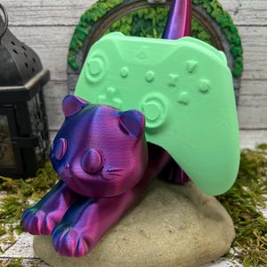 Può includere: Una statuetta di gatto stampata in 3D di colore viola e blu che tiene un controller di videogiochi verde. Il gatto è sdraiato su una roccia verde.